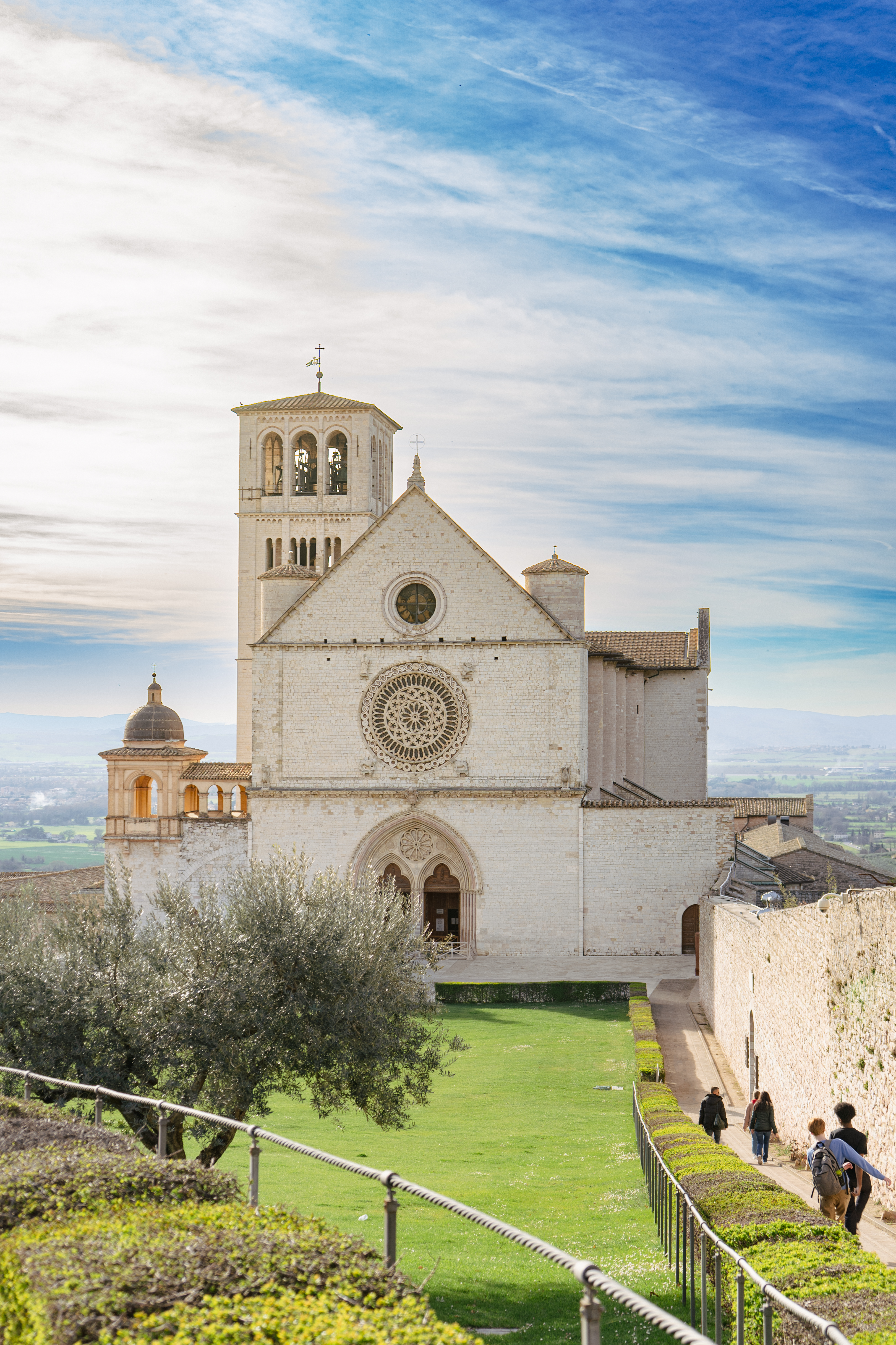 A large, pale stone basilica with a bell tower under a bright sky with people walking on a path.