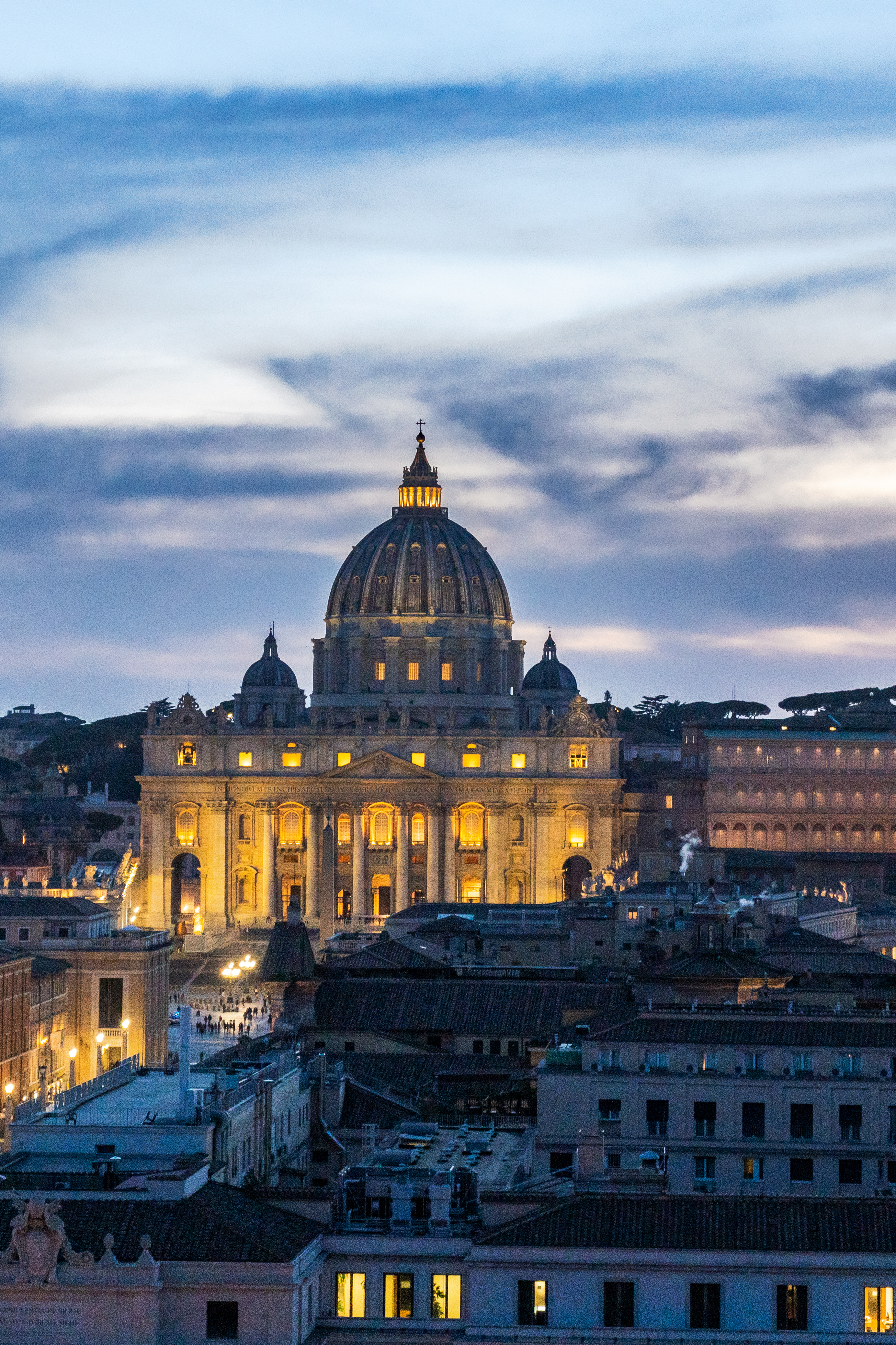 St. Peter's Basilica illuminated against a cloudy twilight sky above the city of Rome.
