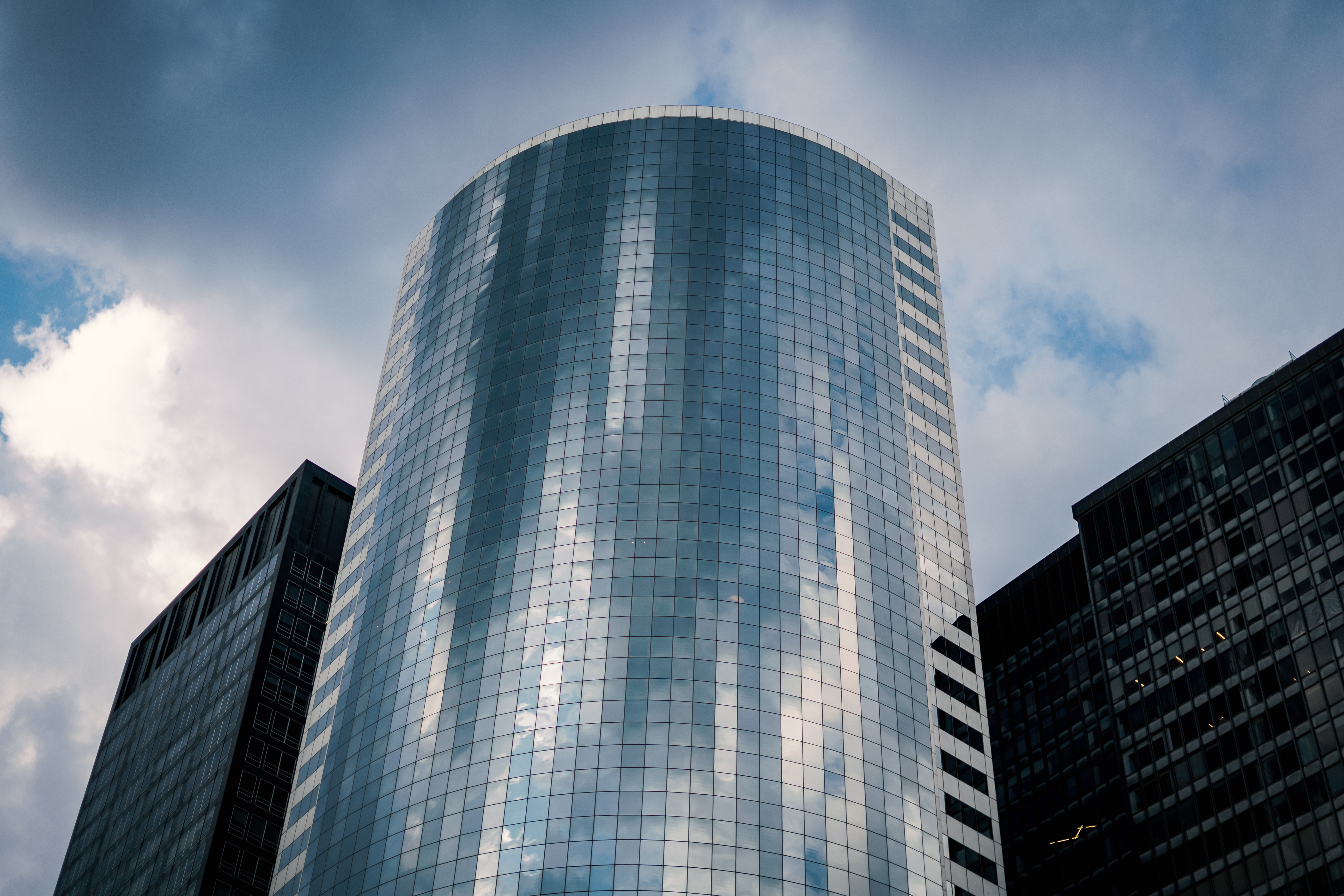 A modern curved glass skyscraper surrounded by darker buildings under a cloudy sky.