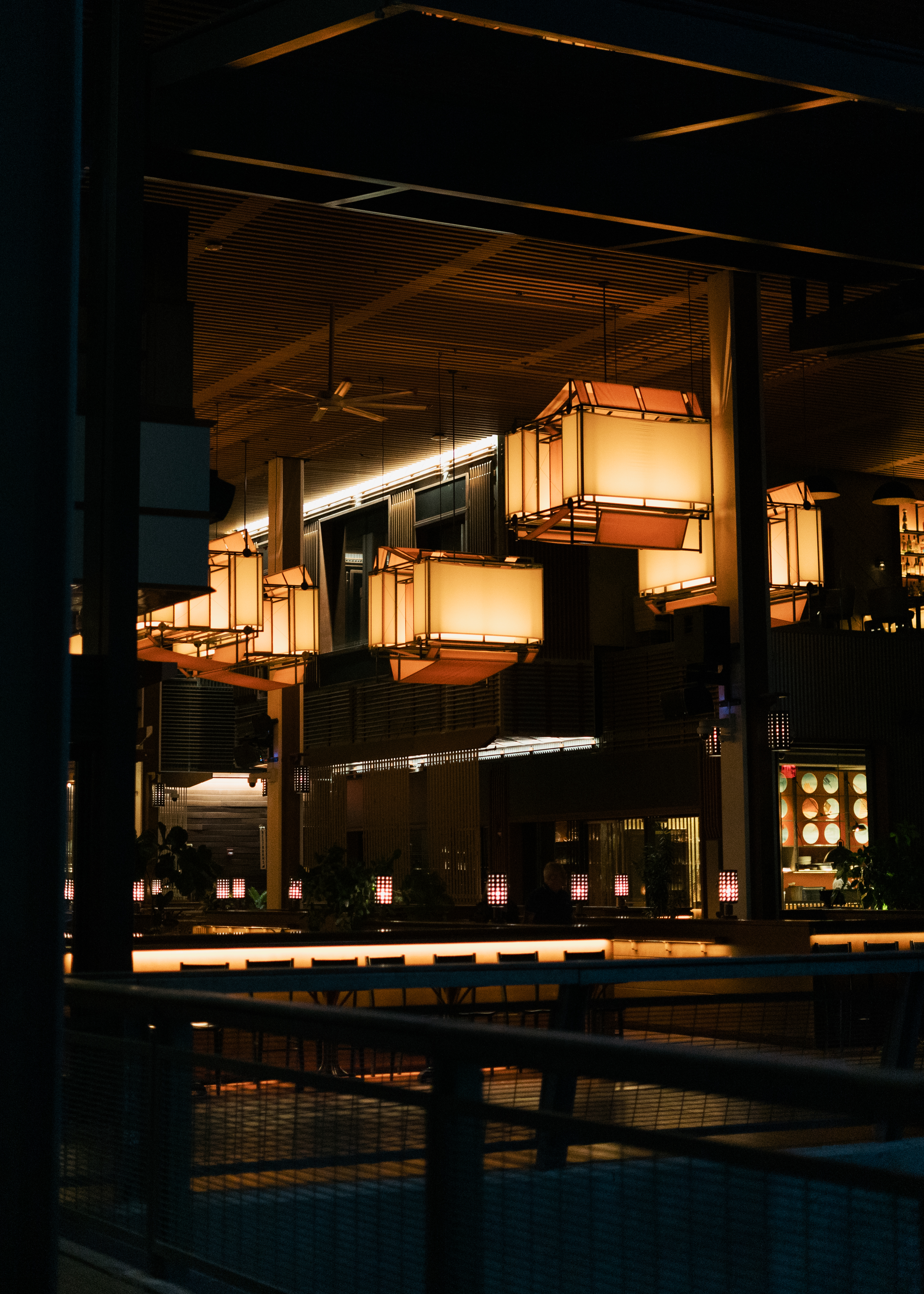 Modern outdoor space at night with hanging box lanterns, a long illuminated bar, and dark framing.