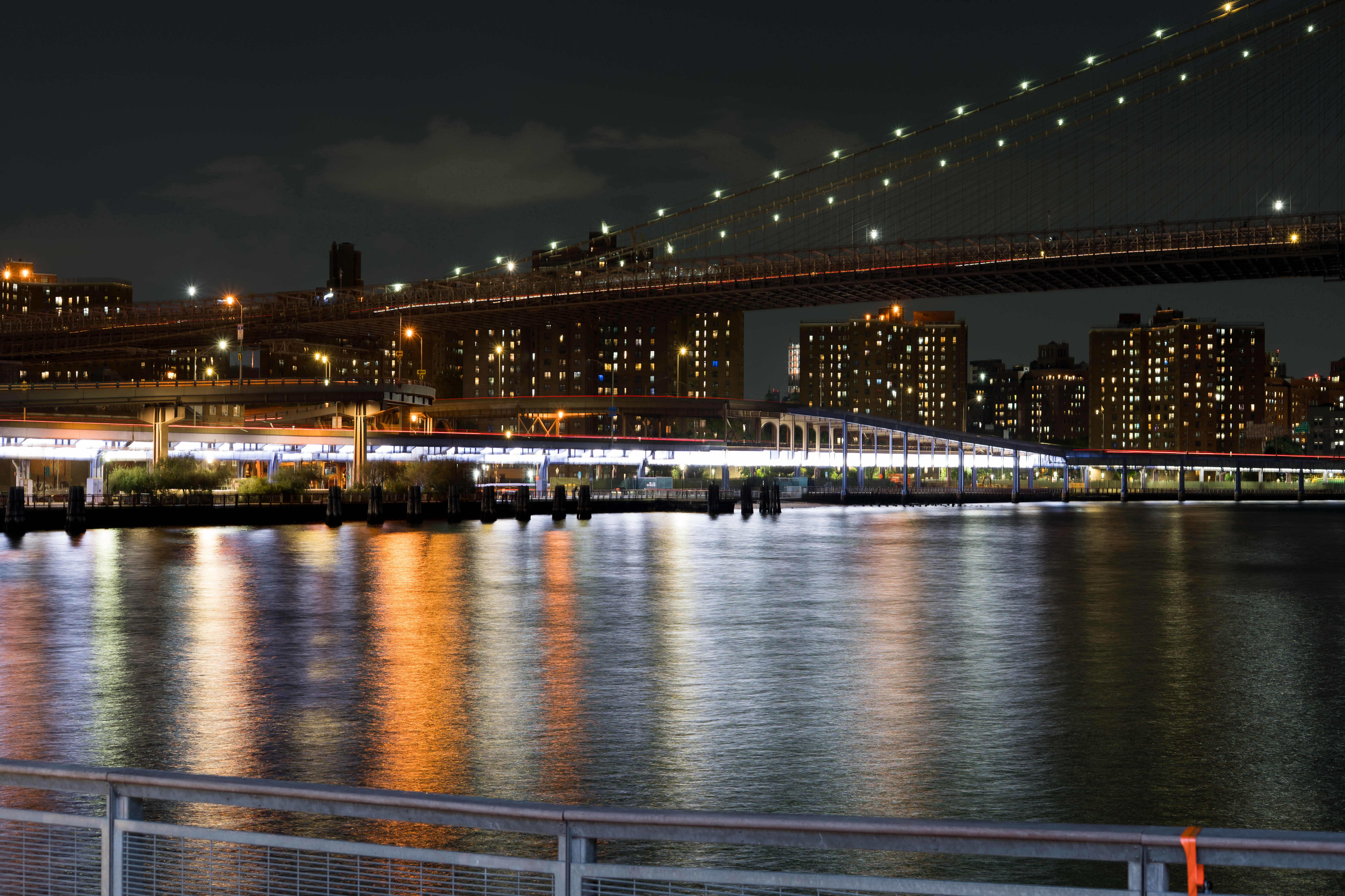 A night view of the Brooklyn Bridge with light streaks and building reflections on the river.